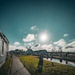 Take a look at this stunning Dutch countryside scene! It’s got windmills, a clear blue sky, and a sunlit pathway. The wooden house on the left adds a cozy touch, while the small canal reflects the sunlight, making it look like a dream come true. And guess what? People are enjoying the scenery, making it a peaceful yet lively place to be.