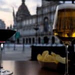 Two Glasses of Wine and Beer on a Table Outside a Building in the Afternoon - Two glasses of wine and beer sit on a table outside a building. The glass of wine is on the left and the glass of beer is on the right. A bowl of chips is in the center of the table. The building behind the table is blurred and out of focus.
